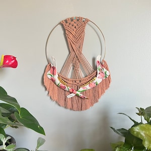May include: A macrame wall hanging with a circular frame and a pink and white floral design. The hanging features a floral pattern with a gold accent.