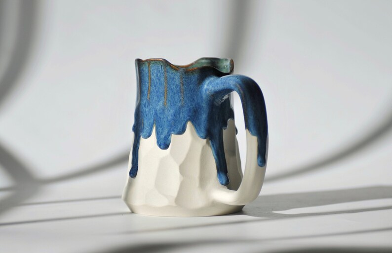 Puede incluir: Una taza de cer&aacute;mica blanca con un esmalte azul que gotea por los lados. La taza tiene una superficie texturizada &uacute;nica y un asa.