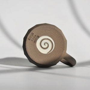 May include: A brown ceramic mug with a white spiral design on the bottom. The mug has the text "GRAM CERA MICSA" on the bottom.