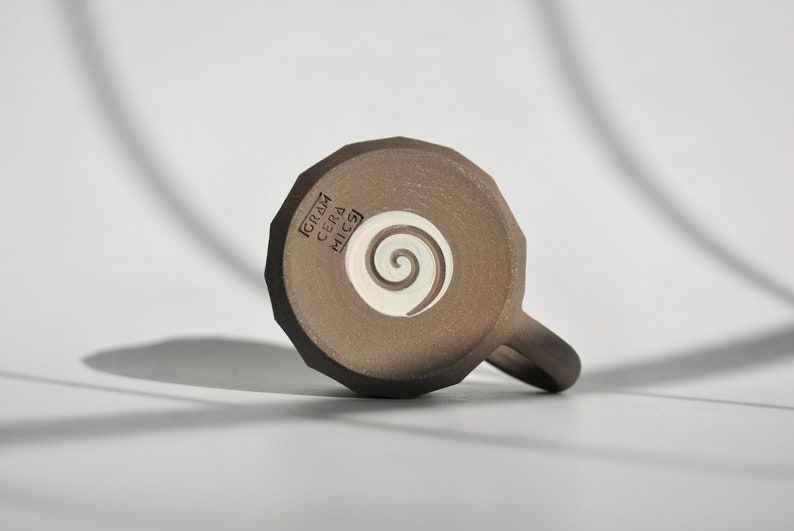 May include: A brown ceramic mug with a white spiral design on the bottom. The mug has the text "GRAM CERA MICSA" on the bottom.