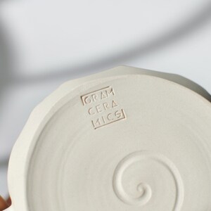 Puede incluir: Un plato de cer&aacute;mica blanco con un dise&ntilde;o en espiral y las palabras "GRAM CERAMICS" estampadas en la parte inferior.