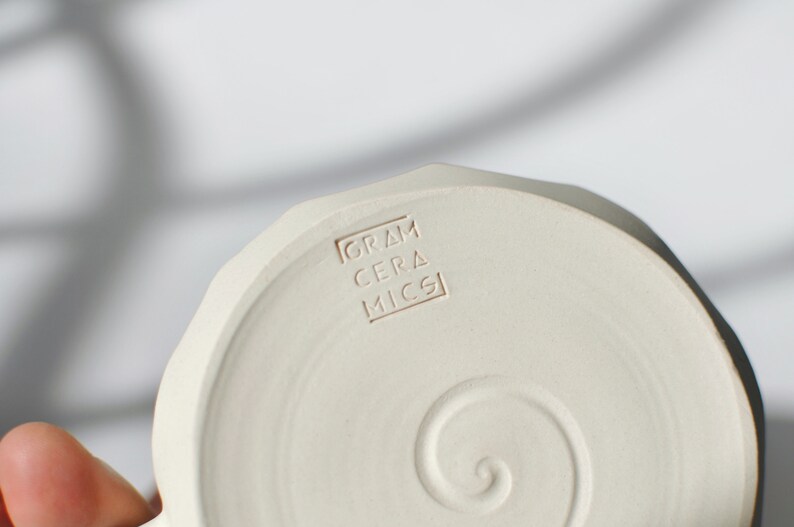 Puede incluir: Un plato de cer&aacute;mica blanco con un dise&ntilde;o en espiral y las palabras "GRAM CERAMICS" estampadas en la parte inferior.
