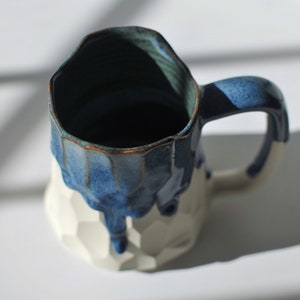 Puede incluir: Una taza de cer&aacute;mica blanca con un esmalte azul que gotea por los lados. La taza tiene un dise&ntilde;o texturizado y geom&eacute;trico.