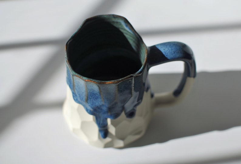 Puede incluir: Una taza de cer&aacute;mica blanca con un esmalte azul que gotea por los lados. La taza tiene un dise&ntilde;o texturizado y geom&eacute;trico.