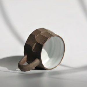May include: A brown ceramic mug with a textured, rocky exterior and a white glazed interior. The mug has a rounded handle and is lying on its side.