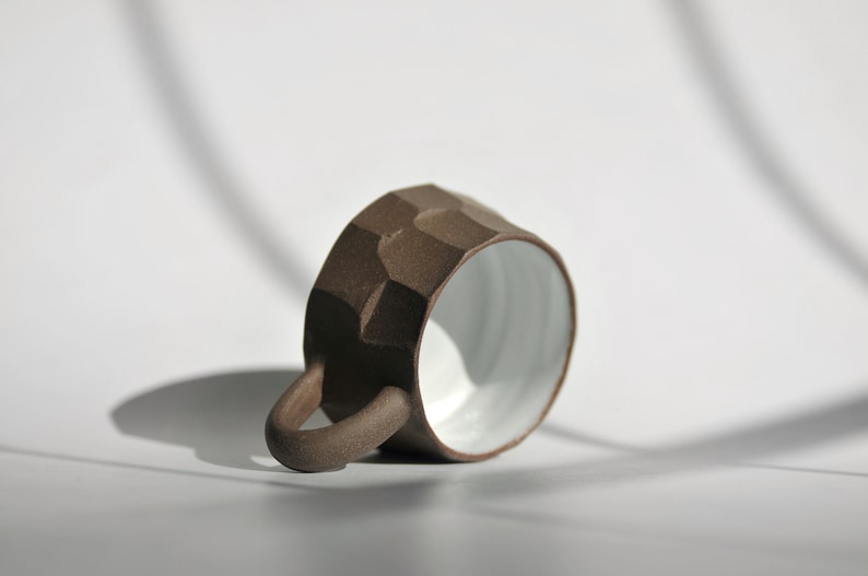 May include: A brown ceramic mug with a textured, rocky exterior and a white glazed interior. The mug has a rounded handle and is lying on its side.