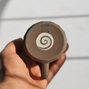 May include: A brown ceramic mug with a white spiral design and the text "GRAM CERA MICSI" on the bottom.