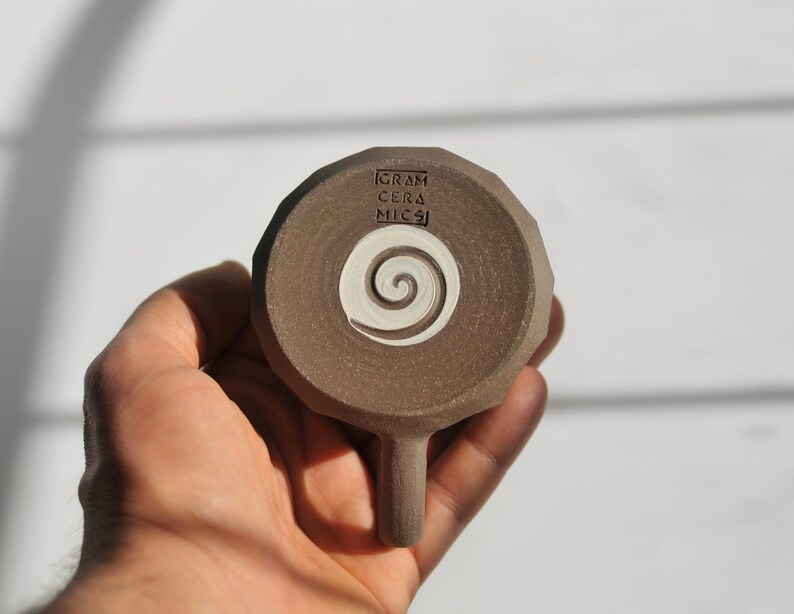 May include: A brown ceramic mug with a white spiral design and the text "GRAM CERA MICSI" on the bottom.