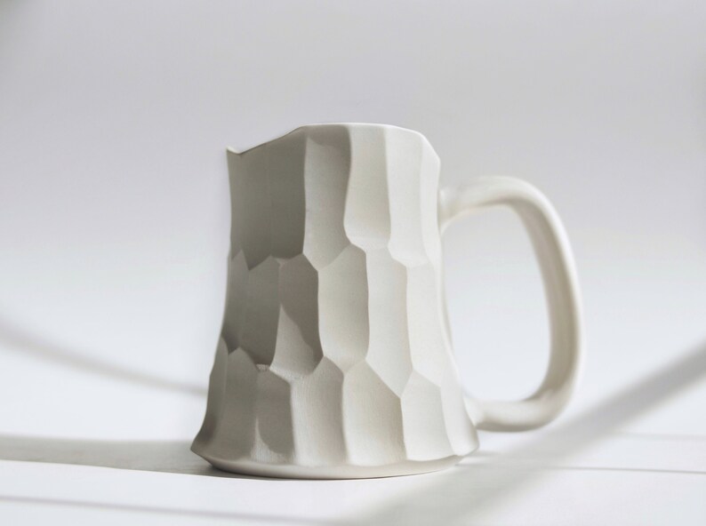 May include: A large and white ceramic mug with a textured, geometric design. The mug has a wide base and a large handle. The mug is on a white surface.