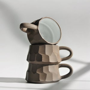 May include: Three stacked ceramic mugs with a textured, brown exterior and a smooth, white interior. The mugs have a rounded shape and a handle on each side.