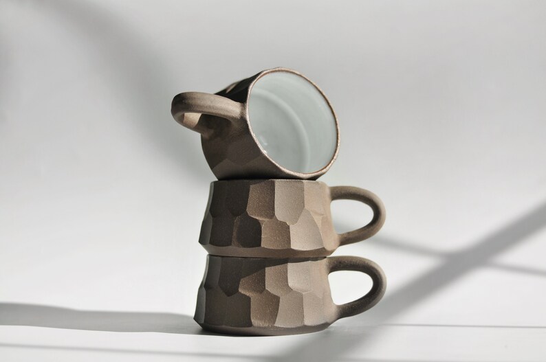 May include: Three stacked ceramic mugs with a textured, brown exterior and a smooth, white interior. The mugs have a rounded shape and a handle on each side.