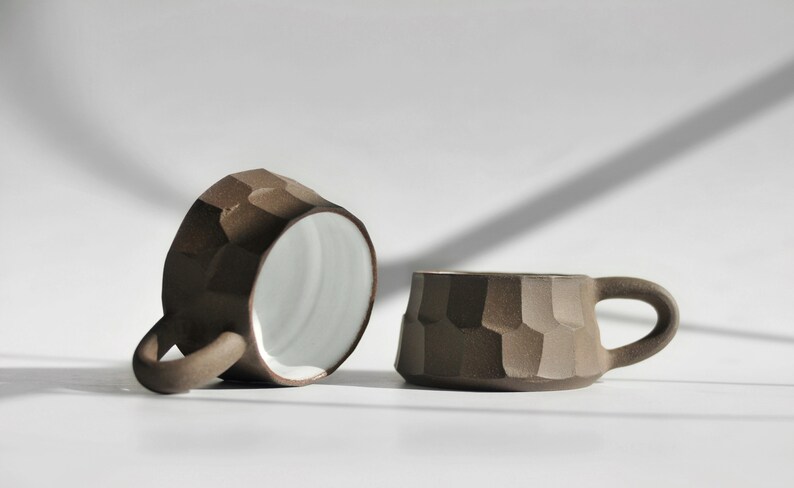 May include: Two brown ceramic mugs with a textured, geometric design. The mugs have white interiors and are on a white surface.
