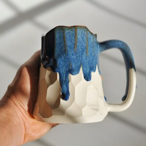 Puede incluir: Una taza de cer&aacute;mica blanca con un esmalte azul que gotea por los lados. La taza tiene un dise&ntilde;o texturizado, similar a un panal de abejas.