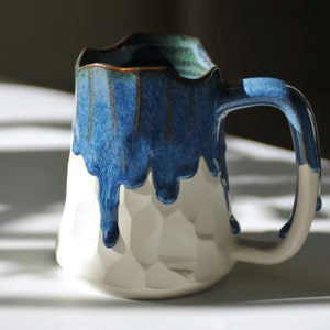 Puede incluir: Una taza de cer&aacute;mica blanca con un esmalte azul que gotea por los lados. La taza tiene una superficie texturizada y un asa.