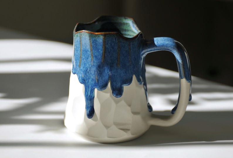 Puede incluir: Una taza de cer&aacute;mica blanca con un esmalte azul que gotea por los lados. La taza tiene una superficie texturizada y un asa.