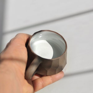 May include: A brown ceramic mug with a white interior and a geometric design. The mug is held in a hand.