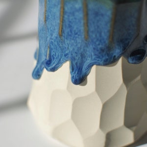 Puede incluir: Un macetero de cer&aacute;mica blanca con un dise&ntilde;o geom&eacute;trico. La parte superior del macetero est&aacute; cubierta con un esmalte azul que gotea por los lados.