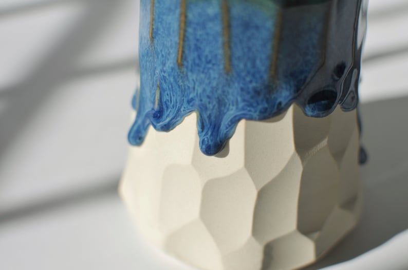 Puede incluir: Un macetero de cer&aacute;mica blanca con un dise&ntilde;o geom&eacute;trico. La parte superior del macetero est&aacute; cubierta con un esmalte azul que gotea por los lados.