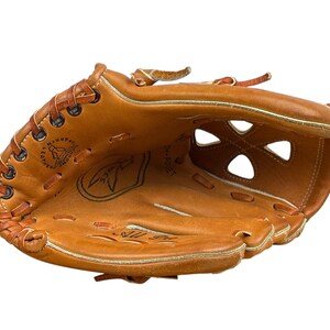 May include: A brown leather baseball glove with a white stitching detail and a circular logo with the text "The Lumberjack" inside. The glove has a webbed design and a lace-up wrist strap.