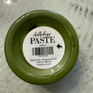 May include: A round olive green container of Chalkology Paste in the color Pesto. The white label features the text Chalkology PASTE, PESTO, a barcode, P193108, Made in USA, 3 US fl oz, 88.7 ml, and chalkcouture.com with a black arrow icon.