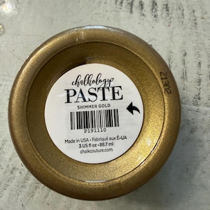 May include: A round container of Chalkology Paste in Shimmer Gold. The white label features black text reading Chalkology PASTE SHIMMER GOLD, a barcode, and the volume 3 US fl oz. The container has a metallic gold finish and a circular lid.