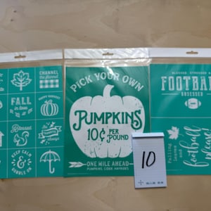 May include: Three teal and white adhesive silk screen stencils in clear plastic sleeves. The left sheet has twelve small fall-themed designs. The center sheet features a large pumpkin graphic with text reading PICK YOUR OWN PUMPKINS 10 CENTS PER POUND. The right sheet displays football-themed text and graphics. A small white notepad with the number 10 written on it is placed in front.