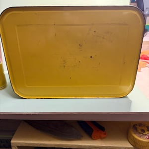 May include: A rectangular metal tray with a bright yellow finish and rounded corners. The surface features a raised border and shows signs of wear, including minor scratches and small dark spots across the central area of the tray.