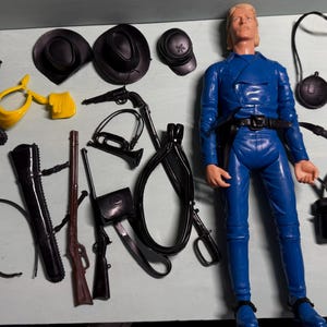 May include: A blue action figure with blonde hair, wearing a blue jumpsuit, boots, and belt. Accessories include black hats, holsters, guns, and a yellow object. A vintage toy set.