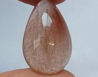 May include: A teardrop-shaped gemstone with a translucent, reddish-brown hue. The stone has internal veining and a reflective, polished surface. The gem is held between two fingers, showcasing its size and shape. The top of the image reads "FIREFLY GEMS AND JEWELLERY".