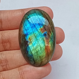 May include: An oval-shaped labradorite gemstone with a vibrant display of blue, green, and orange iridescence. The polished stone is held in a hand, showcasing its natural beauty and unique color play. The gemstone is a focal point.