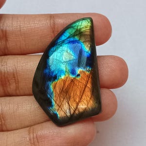 May include: A polished labradorite gemstone with a dark base color and iridescent flashes of blue, teal, and orange. The stone has a smooth, curved shape and is held in a hand. The text "FIREFLY GEMS AND JEWELLERY" is at the top.