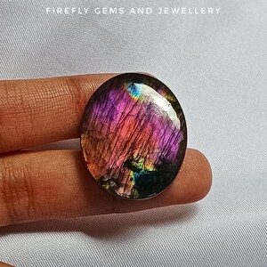 May include: A round, iridescent gemstone cabochon with a purple, pink, and green colour pattern. The gemstone has a smooth, polished surface.