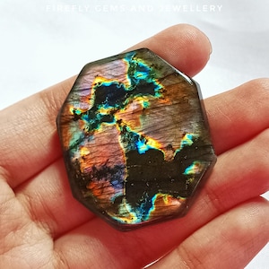 May include: A faceted, iridescent labradorite gemstone with a mix of colors including blue, orange, and green. The stone has a dark base with colorful flashes. The top of the image reads "FIREFLY GEMS AND JEWELLERY".
