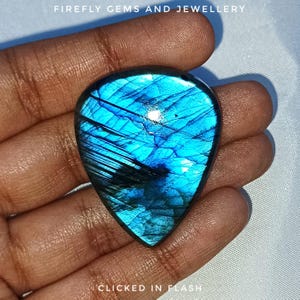 May include: A pear-shaped, shimmering blue gemstone with dark markings. The stone is held in a hand, with the text "FIREFLY GEMS AND JEWELLERY" and "CLICKED IN FLASH" visible. The gemstone has a polished surface.