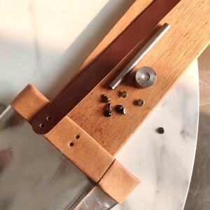 May include: A close-up of a leather craft project with tools and supplies. A brown leather strap is attached to a silver and brown leather bag. The tools include a metal punch, a metal rod, and several metal rivets.