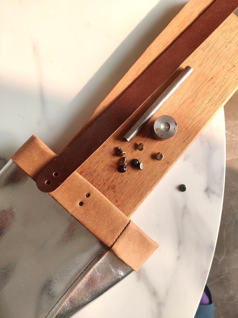 May include: A close-up of a leather craft project with tools and supplies. A brown leather strap is attached to a silver and brown leather bag. The tools include a metal punch, a metal rod, and several metal rivets.
