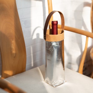 May include: A silver wine bottle carrier with a brown leather handle and a red wine bottle inside. The carrier is sitting on a white chair.