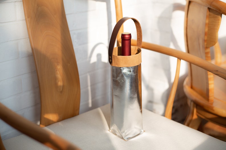 May include: A silver wine bottle carrier with a brown leather handle and a red wine bottle inside. The carrier is sitting on a white chair.