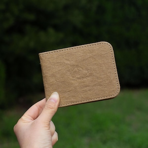 May include: A tan, bi-fold wallet with a textured surface is held in a hand. The wallet has a stitched border and a subtle embossed logo. The background is a blurred green.
