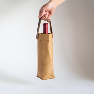 May include: A brown paper wine bottle carrier with a leather handle. The carrier is holding a red wine bottle.