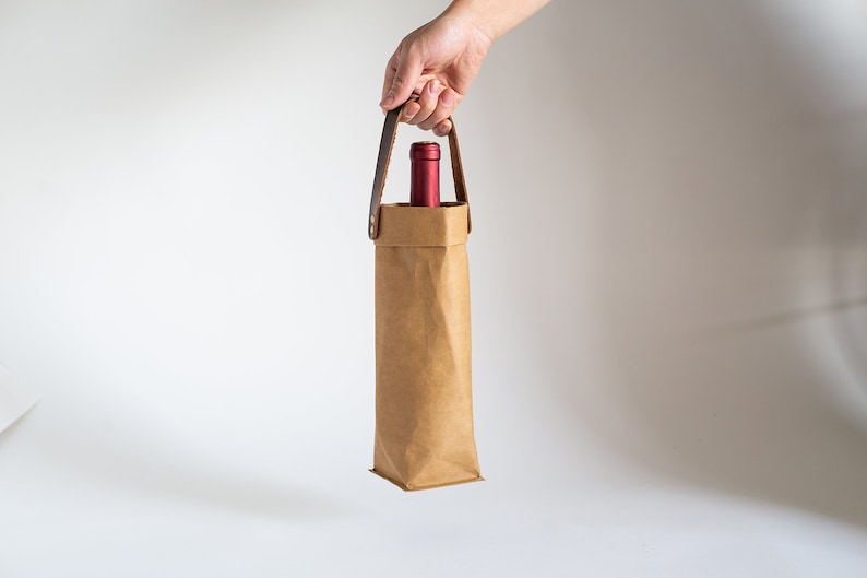 May include: A brown paper wine bottle carrier with a leather handle. The carrier is holding a red wine bottle.