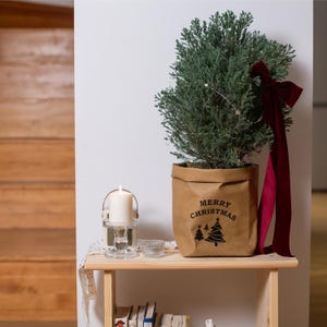 May include: A small evergreen tree in a brown paper pot with "Merry Christmas" printed on it. A burgundy ribbon is tied to the tree. A lit candle sits in a glass holder on a wooden shelf, creating a festive holiday display.