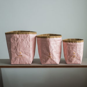 May include: Three pink paper bags with brown paper rims and gold lettering. The bags are crumpled and have the words "organised nature" printed on them.