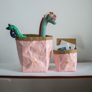 May include: Two pink paper bags with gold lettering. The larger bag has a green and blue stuffed giraffe toy sticking out of the top. The smaller bag has a stack of white cards inside.