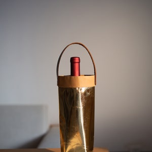 May include: A gold-colored wine bottle carrier with a brown paper liner and a leather handle. The carrier is sitting on a woven placemat.