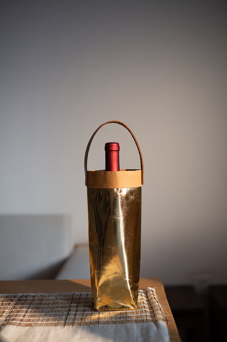 May include: A gold-colored wine bottle carrier with a brown paper liner and a leather handle. The carrier is sitting on a woven placemat.