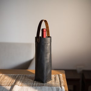 May include: A black leather wine bottle carrier with a brown leather handle. The carrier is sitting on a wooden table with a woven placemat.