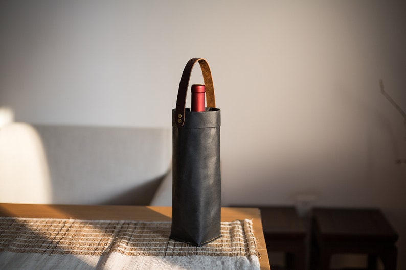 May include: A black leather wine bottle carrier with a brown leather handle. The carrier is sitting on a wooden table with a woven placemat.