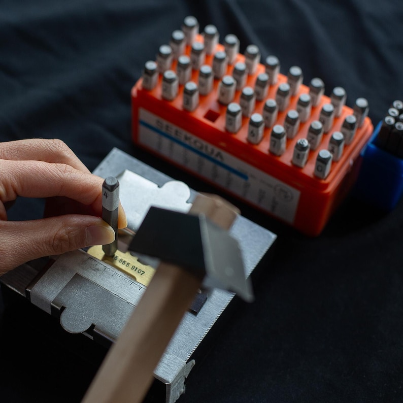 May include: A person using a hammer and punch to stamp metal. The image shows a metal plate with numbers and a hammer. An orange case of bits is in the background.