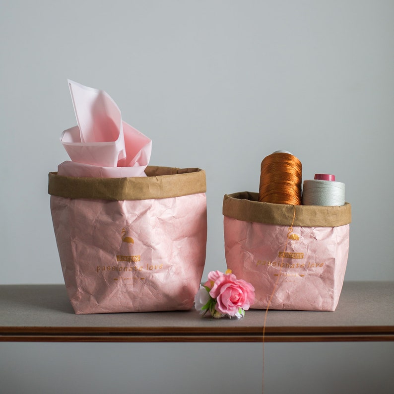 May include: Two pink paper bags with brown tops. The bag on the left has the text "passionate love" printed on it. The bag on the right has the text "passionate love" printed on it. A pink flower is in front of the bags.